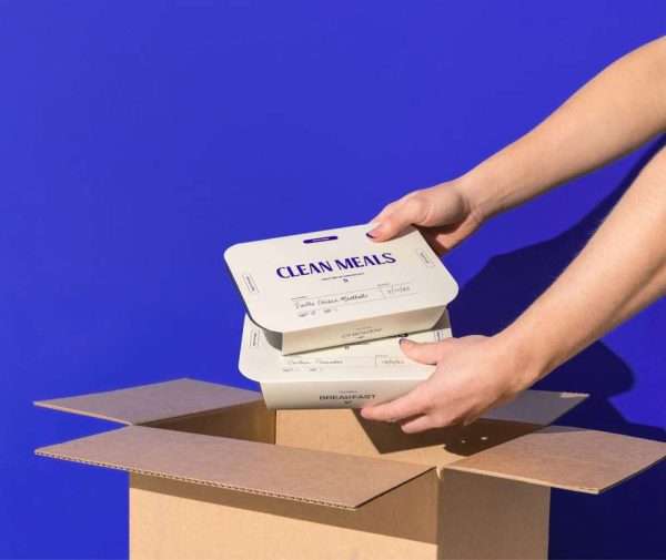 Hands placing 'Clean Meals' packages into a cardboard box against a blue background